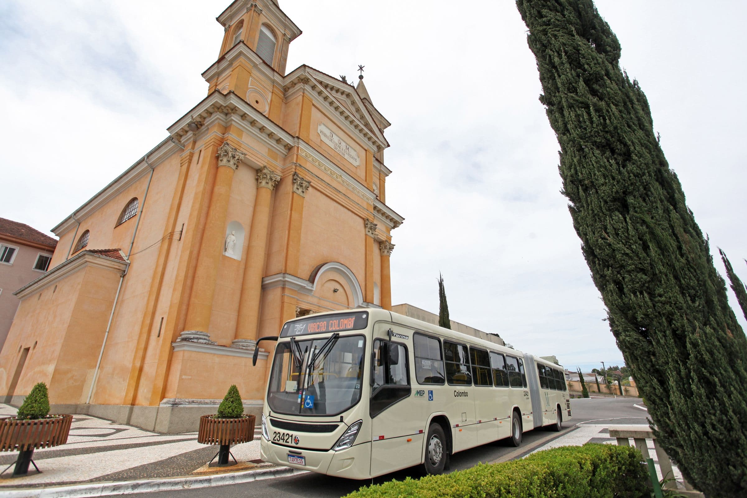 Ônibus articulado diante da Igreja Matriz de Colombo e de um cipreste