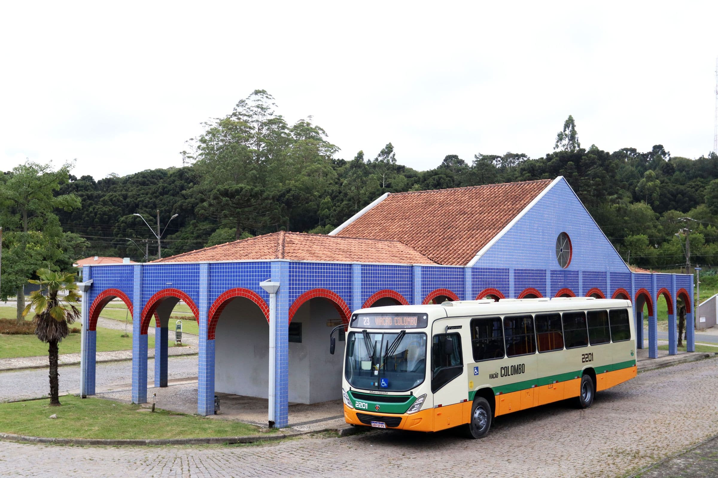 Ônibus da Viação Colombo ao lado de um prédio de arcos, com mata ao fundo