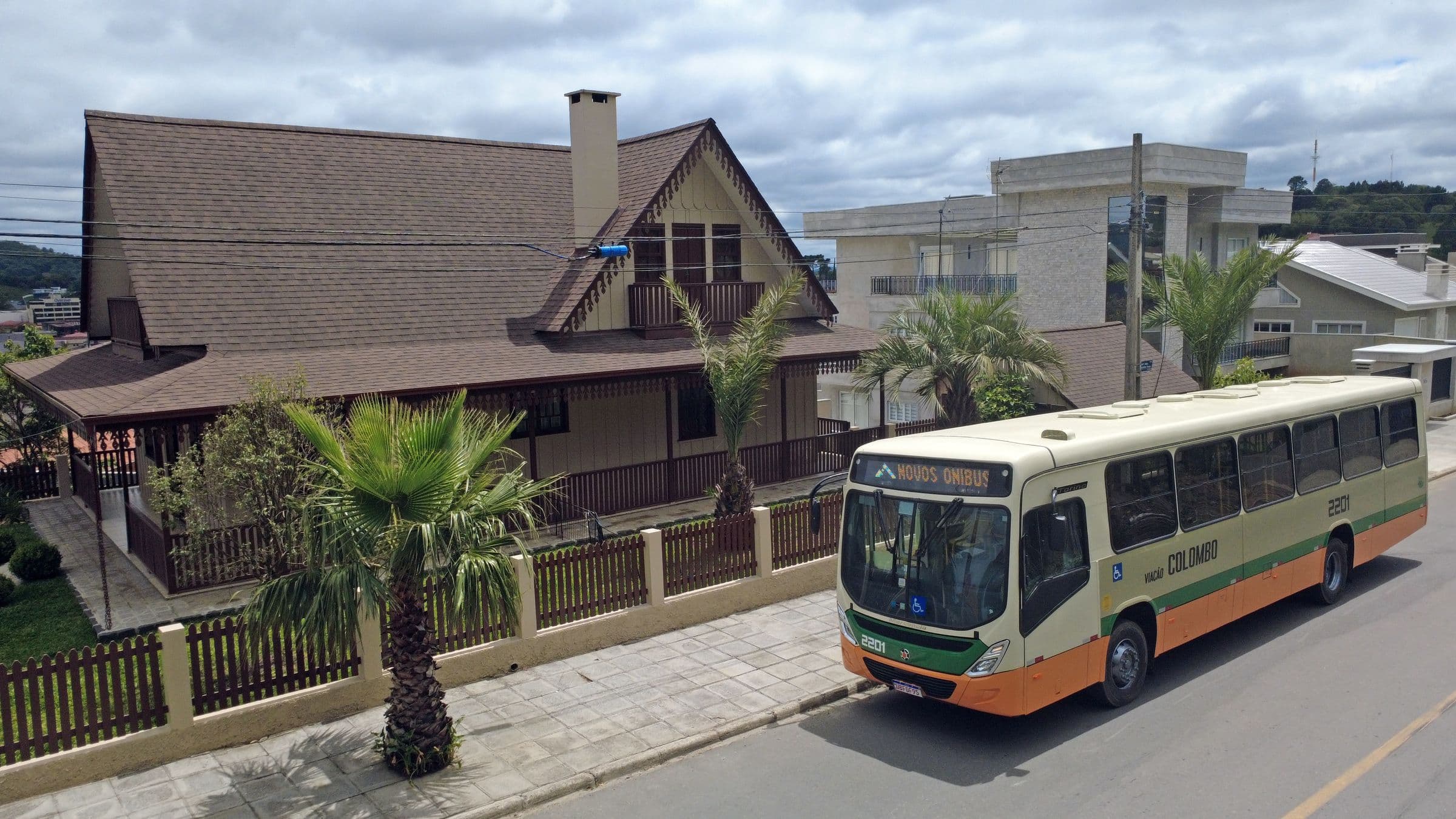 Ônibus da Viação Colombo passando por uma rua arborizada, em frente a uma casa de madeira
